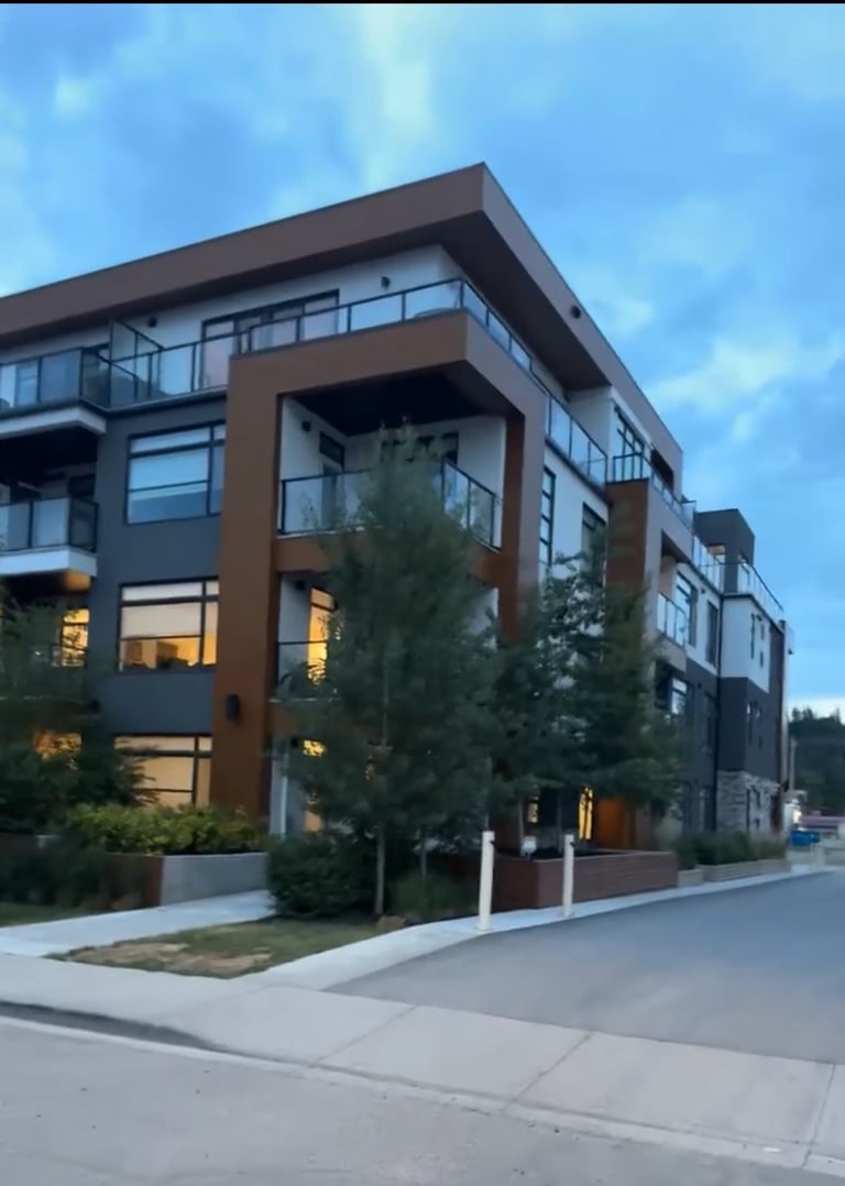 Modern multi-story apartment building with brown and white exterior, large windows, and landscaping, photographed at dusk