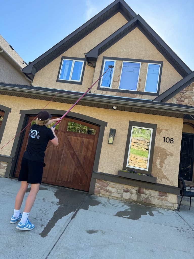 Person cleaning the exterior of a two-story beige and brown house with a pressure washer on the driveway