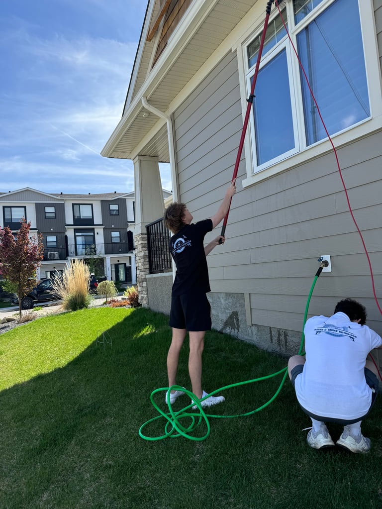 Two people cleaning house exterior with water hose, one standing on green lawn holding red rope, another crouching nearby with green hose coiled on grass