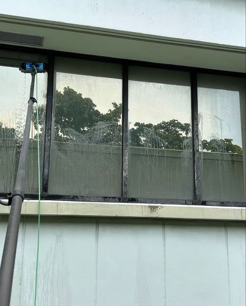 Exterior view of a modern building with three large windows showing trees and sky reflections, with a cleaning equipment pole visible on the left side