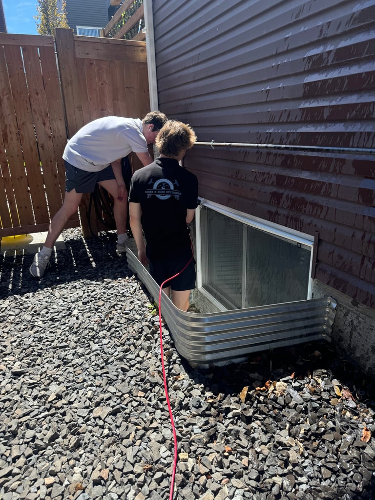 Two people inspecting a metal basement window well in a backyard with gravel ground and wooden fence