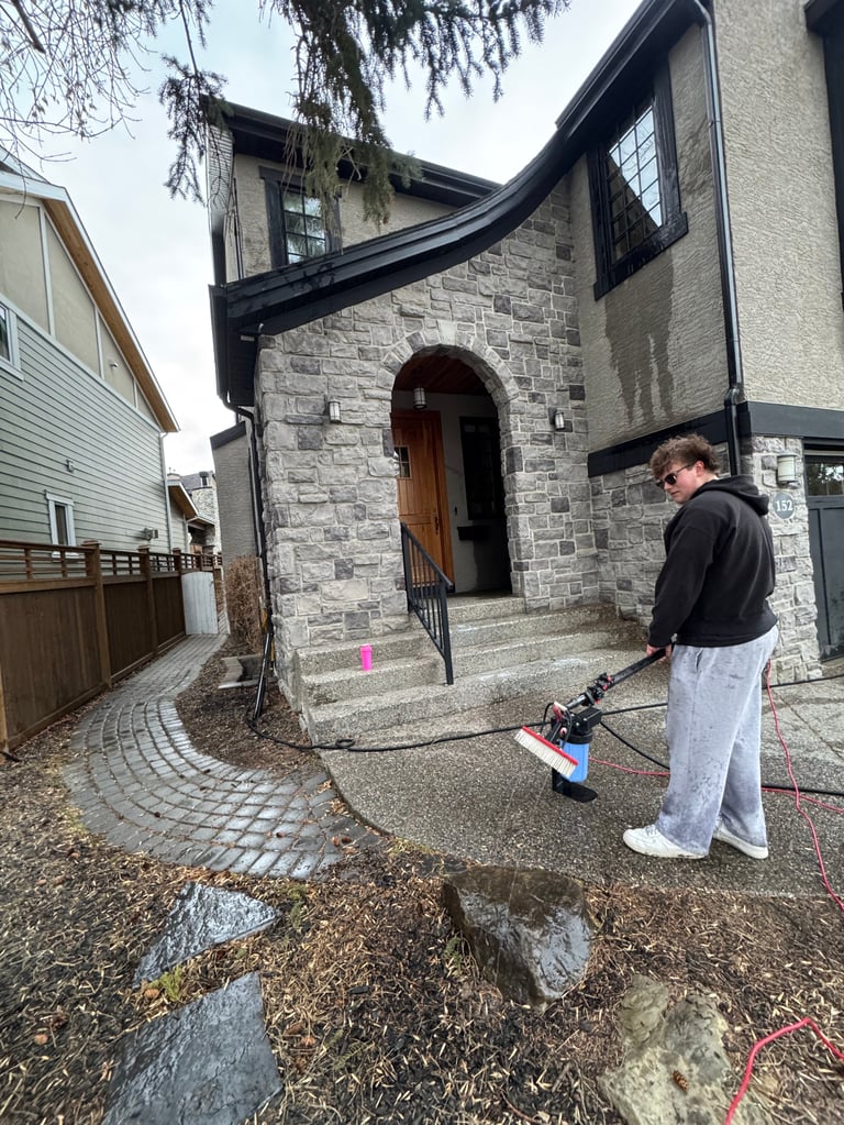 Person using a water hose to clean wet ground in front of a modern stone house with arched black trim
