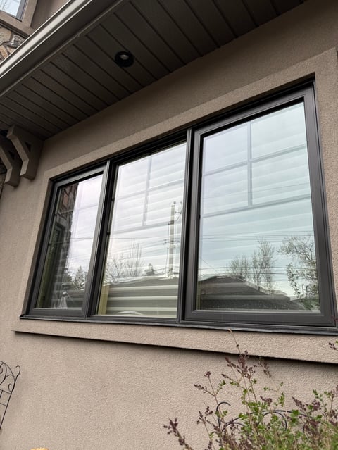 Three large black-framed windows on beige exterior wall with soffited overhang and dried vegetation below