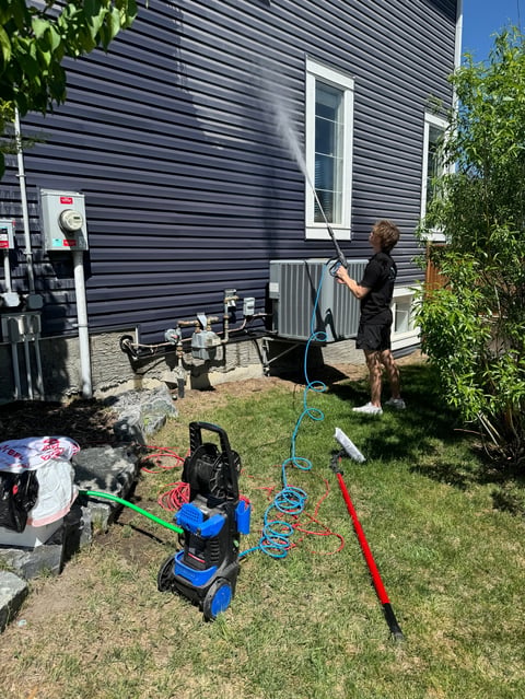 Person power washing a dark siding house exterior with a blue pressure washer equipment in the yard