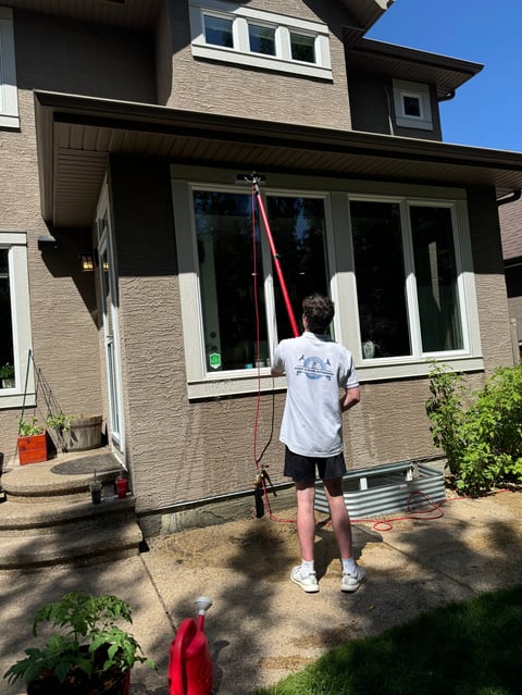 Person in white shirt and shorts standing on patio, raising a red rope or pole toward a residential brick house with windows