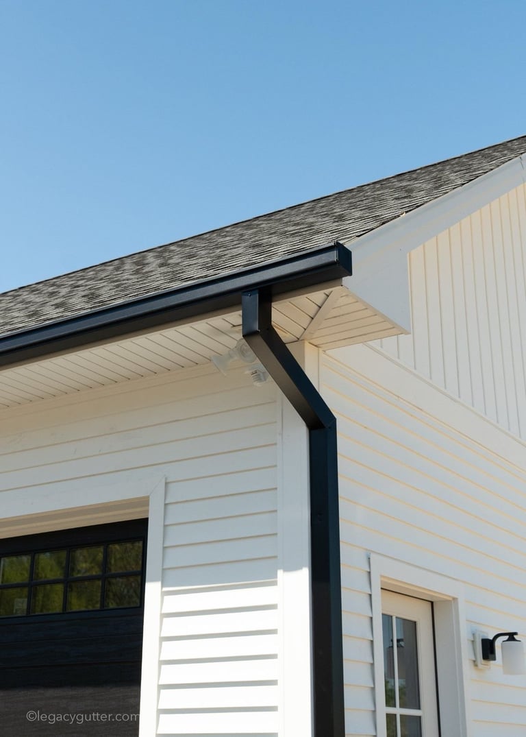 Modern house exterior showing white vinyl siding, black downspout, asphalt shingle roof, and garage door on clear blue sky day